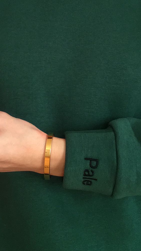 A close-crop onto a woman's arm and wrist. She's wearing a high-quality forest green sweater with the brand name "Pale" embroidered on the wrist cuff.