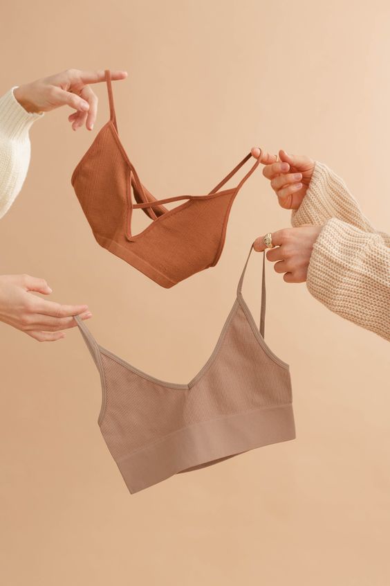 Two sets of women's hands hold up two sport's bras in muted colors against a pale beige backdrop.